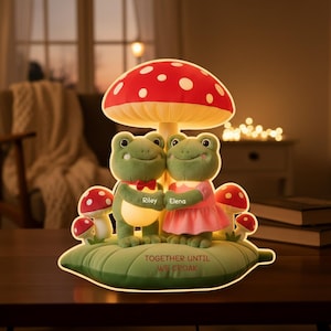 Peut inclure: Une lampe fantaisiste avec deux figurines de grenouilles vertes, l'une avec un nœud papillon et l'autre dans une robe rose, sous un abat-jour en forme de champignon rouge et blanc. La base indique "TOGETHER UNTIL WE CROAK".
