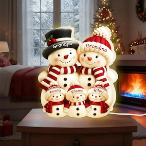 Op de afbeelding: Een kerstversiering van een sneeuwmanfamilie. De sneeuwmannen dragen hoeden en sjaals, met de namen "Grandpa", "Grandma", "Amelia", "Elena" en "Riley" erop. De decoratie is verlicht en staat op een houten tafel.