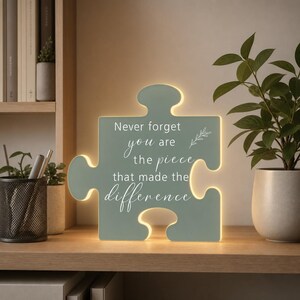 May include: A sage green puzzle piece-shaped LED light with a white printed quote that reads, "Never forget you are the piece that made the difference," accompanied by a small leaf illustration. The light emits a soft glow from its edges.