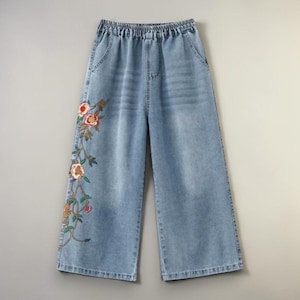 Peut inclure: Pantalon large en jean bleu clair avec une taille élastique. Le pantalon présente une broderie florale aux couleurs rouge, orange et verte le long de la jambe gauche. Il possède deux poches avant.