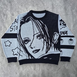 May include: A light blue and black sweater with a manga-style illustration of a woman's face. The sweater has the word "NANA" on the sleeve, along with strawberry and star graphics. The sweater has a crew neck and long sleeves.