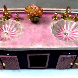 May include: A decorative double sink vanity with a pink countertop and two shell-shaped sinks. The vanity has gold-colored faucets and drawer pulls, and a dark base with decorative doors. A flower sits between the sinks.
