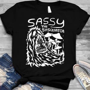 Peut inclure: T-shirt noir avec un dessin blanc d'un Sasquatch et de champignons. Le texte "SASSY the Sasquatch" est au-dessus du dessin. Le t-shirt est fait d'une matière douce, adaptée aux tenues décontractées.