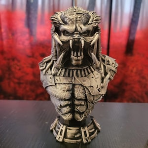 May include: A detailed silver-colored bust of the Predator alien, featuring intricate armor and facial details. The sculpture showcases the creature's iconic mandibles and dreadlocks, with a textured finish. The bust is displayed on a dark surface.