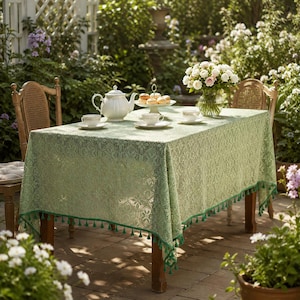 May include: A rectangular sage green lace tablecloth with an intricate floral pattern and a decorative fringe trim along the edges. The tablecloth is draped over a wooden table, featuring a white ceramic teapot, teacups, and a tiered serving stand.