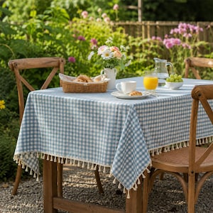 May include: A rectangular table covered with a light blue and white gingham tablecloth featuring a decorative fringe trim along the edges. The table is set with a woven basket of bread, a white ceramic pitcher of flowers, a mug, and a glass of juice.