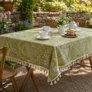 May include: A rectangular olive green tablecloth featuring a white botanical fern and leaf pattern with a decorative fringe trim along the edges. The tablecloth is set with a white ceramic teacup, a saucer, and a plate holding a slice of cake.