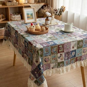 Könnte beinhalten: Ein rechteckiges Tischtuch mit einem Patchwork-Design in Blau-, Lila-, Grün- und Beigetönen. Das Tischtuch hat eine Spitzenborte an den Rändern und liegt auf einem Holztisch. Ein kleines Tablett mit Gebäck und ein gerahmtes Bild befinden sich auf dem Tisch.