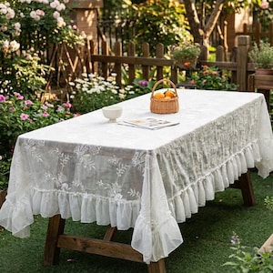 K&ouml;nnte beinhalten: Eine wei&szlig;e rechteckige Tischdecke mit floraler Stickerei und R&uuml;schenkante, drapiert &uuml;ber einem Picknicktisch aus Holz. Ein kleiner Korb mit Orangen und eine wei&szlig;e Sch&uuml;ssel stehen auf dem Tisch in einem Garten.