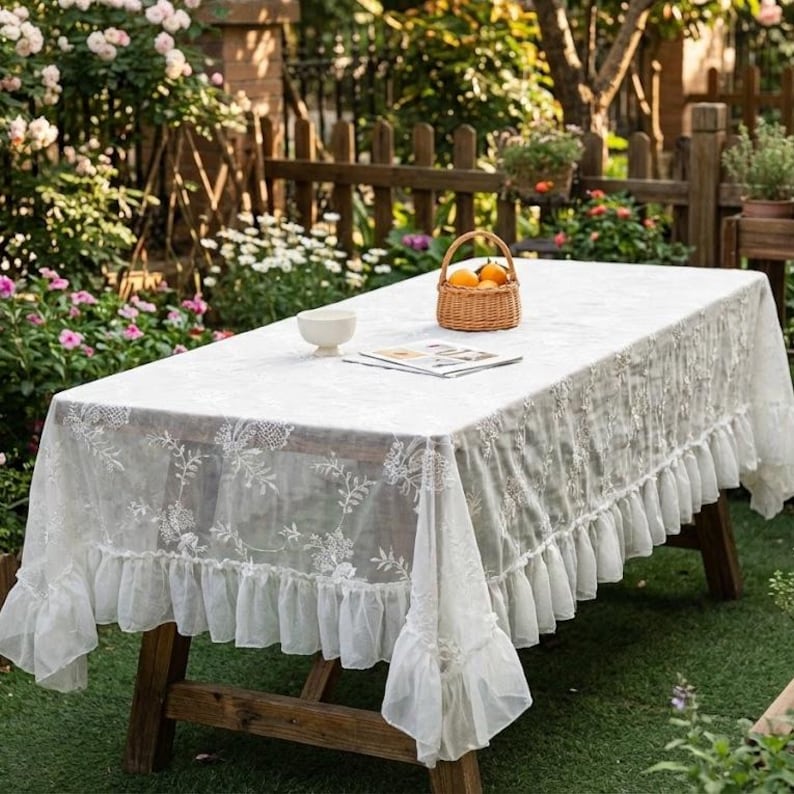 K&ouml;nnte beinhalten: Eine wei&szlig;e rechteckige Tischdecke mit floraler Stickerei und R&uuml;schenkante, drapiert &uuml;ber einem Picknicktisch aus Holz. Ein kleiner Korb mit Orangen und eine wei&szlig;e Sch&uuml;ssel stehen auf dem Tisch in einem Garten.