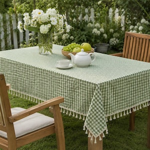 May include: A rectangular tablecloth featuring a green and white gingham check pattern with cream-coloured tassels along the edges. The tablecloth is draped over a table, holding a glass vase of white flowers, a white teapot, and a bowl of fruit.
