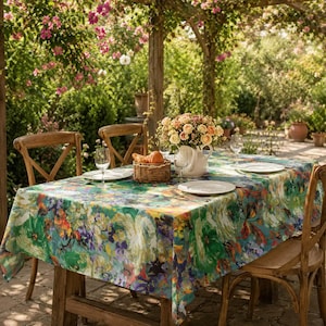 May include: A rectangular tablecloth featuring a vibrant, painterly floral pattern in shades of green, blue, yellow, and orange. The cloth covers a wooden table set with white plates, clear wine glasses, and a central woven basket of fruit.