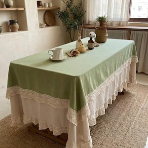 May include: A sage green tablecloth with a white lace trim and a white underskirt. A white teacup and saucer sit on the table, along with decorative items. The table is set on a woven rug.