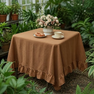 May include: A square, solid brown tablecloth with a ruffled edge draped over a table. On the table sits a white ceramic vase filled with a bouquet of white and pink roses, a small white plate with a round pastry, and a white ceramic teacup.