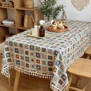 Puede incluir: Un mantel rectangular con un diseño de patchwork colorido, con cuadrados en tonos de azul, verde, naranja y crema. El mantel tiene un borde de flecos blancos y está sobre una mesa de madera. Hay objetos decorativos en la mesa.