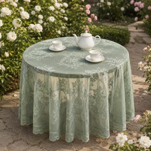Peut inclure: Une table ronde recouverte d'une nappe en dentelle vert sauge ornée d'un motif floral complexe. Sur la table se trouvent une théière en porcelaine blanche et deux tasses à thé assorties avec soucoupes, disposées pour le service du thé.