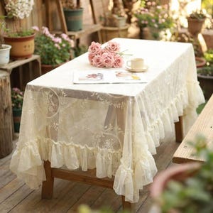 K&ouml;nnte beinhalten: Eine cremefarbene Spitzentischdecke mit R&uuml;schensaum bedeckt einen Holztisch. Die Tischdecke hat Blumen- und Bl&auml;ttermuster. Ein Strau&szlig; rosa Rosen, eine Tasse und eine Untertasse liegen auf dem Tisch.