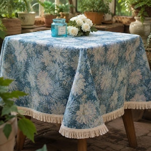 May include: A square tablecloth featuring a blue and white floral pattern with a repeating chrysanthemum design. The fabric has a textured appearance and is finished with a cream-colored fringe trim along all four edges of the square table.