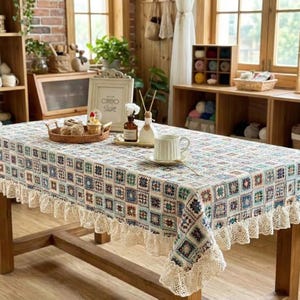 May include: A rectangular tablecloth with a colourful patchwork design, featuring squares of blue, green, and beige. The tablecloth has a cream-coloured lace trim and is set on a wooden table. A cup and saucer are on the table.