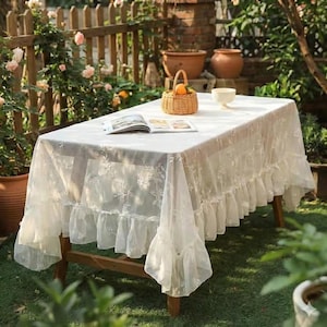 K&ouml;nnte beinhalten: Ein rechteckiger Holztisch, bedeckt mit einer wei&szlig;en Spitzentischdecke. Die Tischdecke hat eine Blumenstickerei und einen ger&uuml;schten Rand. Ein kleiner Weidenkorb mit Orangen, ein Buch und eine wei&szlig;e Sch&uuml;ssel stehen auf dem Tisch. Der Tisch befindet sich im Freien, umgeben von Gr&uuml;n.