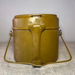 May include: An olive drab metal mess kit with a cylindrical body, a secure locking latch mechanism on the front, and a hinged wire handle attached to the sides. The container features a smooth, painted finish with visible signs of wear.