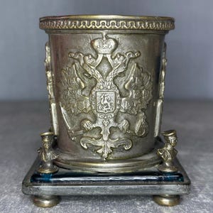 Op de afbeelding: Een zilverkleurige metalen beker met sierlijke details, met een dubbelkoppig adelaarsymbool en decoratieve bustes. De beker staat op een rechthoekige basis met kleine voetjes, met een vintage esthetiek.
