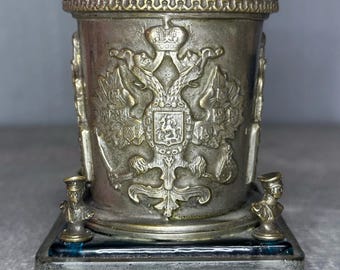 Könnte beinhalten: Ein zylindrisches Schreibtischaccessoire aus Metall mit einem erhabenen Doppeladler-Emblem auf der Vorderseite. Das Gefäß steht auf einem quadratischen Sockel mit kleinen dekorativen Büsten an den Ecken und ruht auf vier abgerundeten Füßen in Metalloptik.