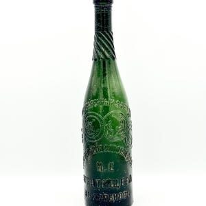 Puede incluir: Una botella de vidrio verde oscuro con un cuello largo y delgado que presenta una textura retorcida. El cuerpo de la botella está grabado con medallones circulares que contienen figuras y texto cirílico, incluyendo las iniciales I.E. y Járkov.