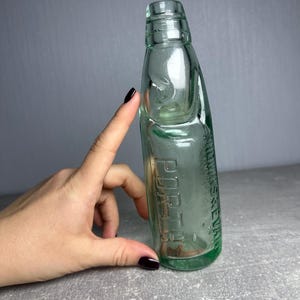 以下が含まれることがあります： 「PORTER」の文字がエンボス加工された、ヴィンテージの薄緑色のガラス瓶。丸みを帯びた形状で、首が細くなっています。手で持っています。