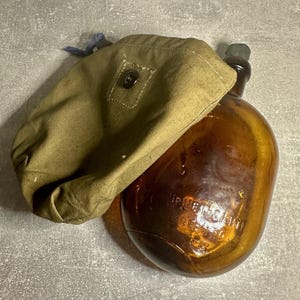 May include: A vintage brown glass canteen with a green cap, partially covered by an olive drab canvas carrying case. The canteen has embossed text. The case has a button closure and a blue drawstring.
