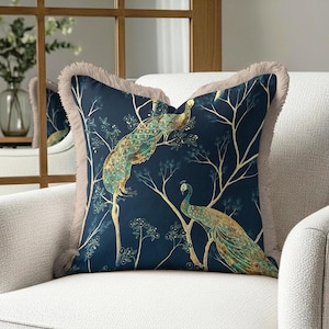 Könnte beinhalten: Dekoratives quadratisches Zierkissen mit marineblauem Hintergrund und einem Pfauenmuster in Gold-, Türkis- und Grüntönen. Das Kissen hat einen Fransenrand und liegt auf einem weißen Sessel. Maße in Zentimetern.