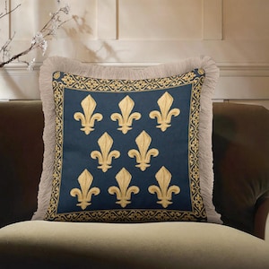 May include: Square decorative pillow with a navy blue center featuring gold fleur-de-lis embroidery. The pillow has a gold patterned border and a beige fringe trim. The pillow is placed on a brown sofa.