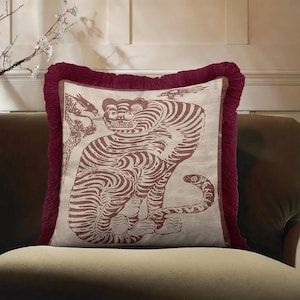 May include: A square throw pillow featuring a beige fabric cover with a dark red printed illustration of a seated tiger. The pillow is framed by a thick, dark red fringe border along all four edges, creating a textured and decorative appearance.