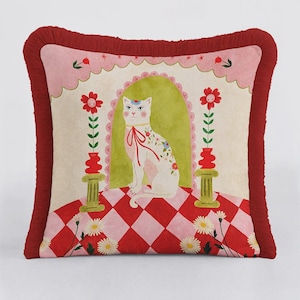 Op de afbeelding: Decoratief sierkussen met een grillige kattenillustratie. De witte kat heeft bloemaccenten en zit op een rood-roze geblokt oppervlak. Het kussen heeft een rode franjeborder en een roze geschulpte bovenrand.