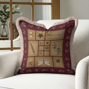 May include: Decorative square pillow with a botanical design. The pillow features a central panel with various plant and insect illustrations in a grid pattern, framed by a burgundy border with floral accents. The pillow has a fringed edge and is placed on a white armchair.