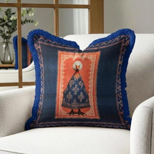 May include: Decorative square pillow with a blue background and a central image of a swan wearing a patterned blue robe. The pillow has a blue fringe border and is placed on a white armchair.