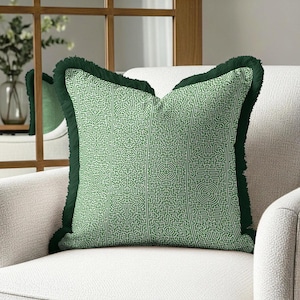 Puede incluir: Cojín decorativo verde con un patrón de laberinto. El cojín tiene un ribete de flecos verde oscuro y está colocado en un sillón blanco. El diseño del cojín presenta un patrón geométrico repetitivo en un tono verde más claro.
