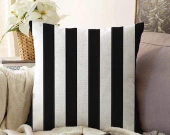 May include: Square throw pillow with a bold black and white striped pattern. The pillow is made of a textured fabric and is placed on a cream-colored blanket. Decorative home accent.