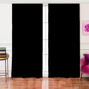 May include: Two solid black window curtains hanging in front of a white window frame. The curtains are made of a thick, opaque material. The curtains are hanging in front of a window with a white frame. The curtains are hanging in front of a window with a white frame.