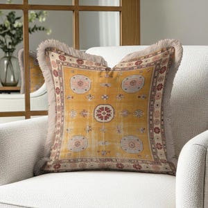 Può includere: Un cuscino quadrato decorativo con sfondo giallo senape e un design dettagliato con motivi nei toni del rosso, crema e beige. Il cuscino ha un bordo con frange ed è posto su una poltrona bianca.
