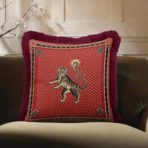 May include: A square throw pillow featuring a red fabric cover with a repeating white geometric pattern. The center displays a tiger and a cobra illustration. The pillow is framed by a black and red border with corner motifs and deep red fringe.