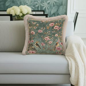 May include: Square cushion with a teal background, featuring pink floral and bird designs. The cushion has a fringed border and is placed on a light grey sofa. A white blanket is draped over the sofa.