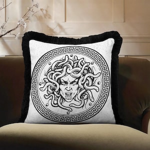 May include: White square throw pillow featuring a black and white Medusa illustration. The Medusa design is centered on the pillow, surrounded by a Greek key border. The pillow has a black fringe border and is placed on a brown sofa.