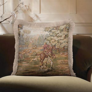 May include: Square decorative pillow with a tapestry design depicting a hunting scene with a rider on horseback, hounds, and a landscape. The pillow has a beige fringe trim and is placed on a green sofa.