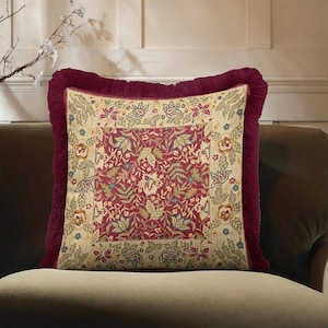May include: Square cushion with a floral design in red, beige, and green. It has a burgundy fringe border and is on a brown sofa. The cushion's pattern includes leaves and flowers, creating a classic look.