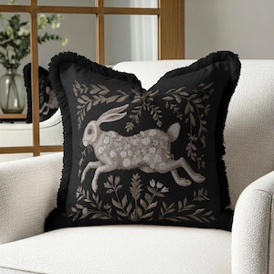May include: Black square throw pillow featuring a running rabbit design. The rabbit is light gray with floral detailing, surrounded by a brown botanical pattern. The pillow has a black fringe border and is placed on a white armchair.