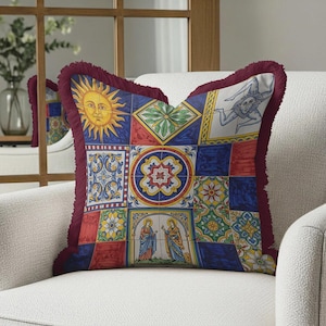 May include: Square decorative pillow with a patchwork design featuring sun, geometric, and religious imagery. The pillow has a burgundy fringe trim and is placed on a white chair. The design incorporates red, blue, yellow, and green colors.