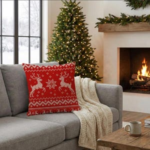 May include: A red knitted throw pillow with a reindeer and snowflake design. The pillow is on a gray sofa with a cream-colored knitted throw blanket. A Christmas tree and fireplace are in the background.