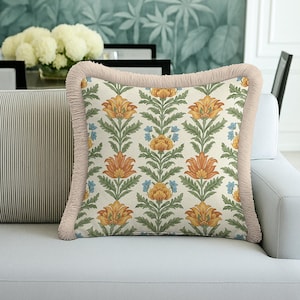 May include: Square decorative pillow with a floral pattern in orange, yellow, green, and blue on an off-white background. The pillow has a light pink fringe trim. The pillow is on a white sofa.