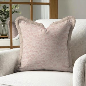 May include: A square decorative pillow with a light pink pattern and a fringed border. The pillow is placed on a white armchair, creating a soft and inviting aesthetic. The pattern appears to be a repeating fan or petal design.